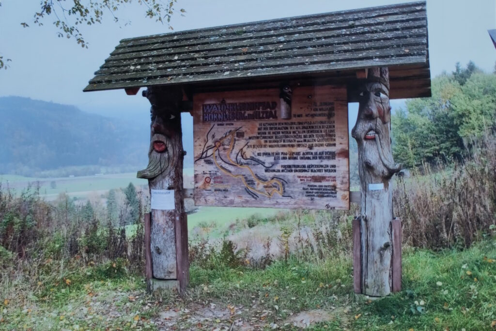 Startportal zum Walderlebnispfad: Auf einer Holztafel ist die Wegstrecke eingezeichnet. Die Tafel ist links und rechts von geschnitzten Waldmännle mit freundlichen Gesichtern flankiert.